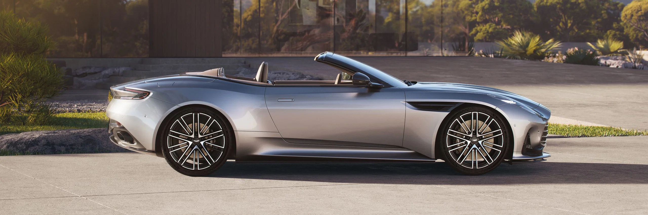 Silver Aston Martin convertible parked in front of a modern glass house.