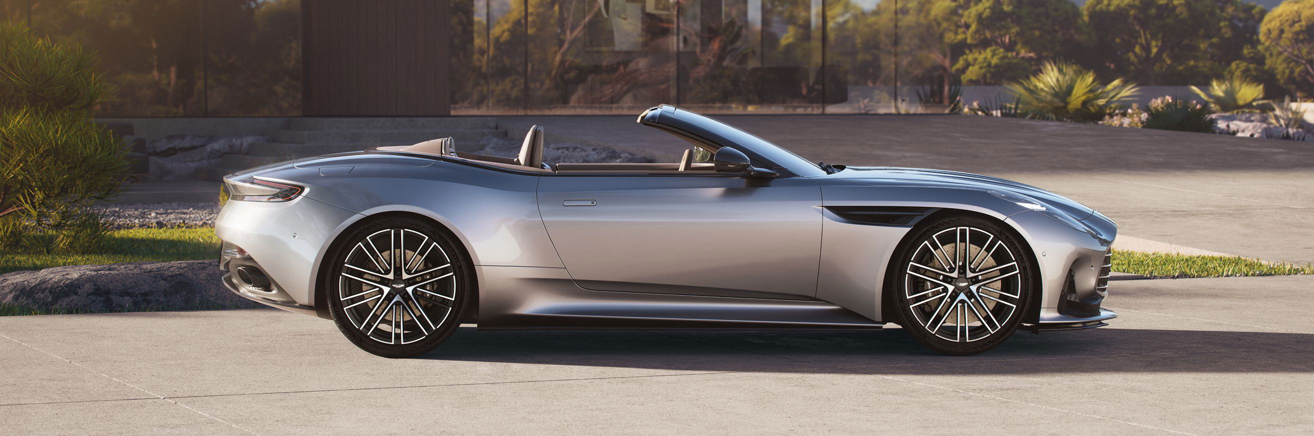Silver Aston Martin convertible parked in front of a modern glass house.